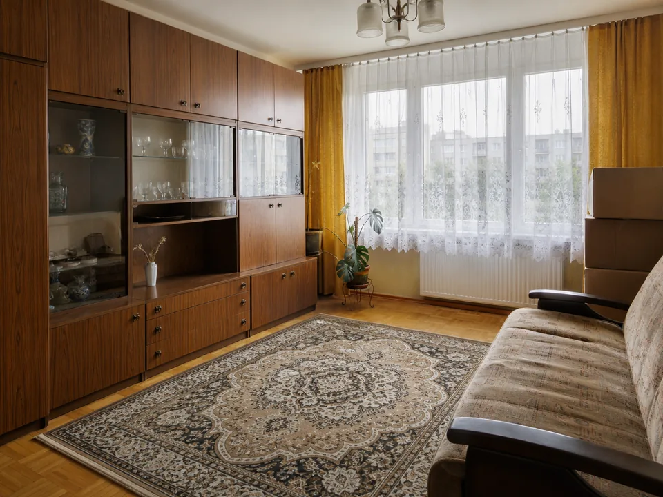 Salon w mieszkaniu w bloku z lat 80. z meblościanką, dywanem i kartonami przy ścianie, przygotowany do decyzji o sprzedaży lub najmie